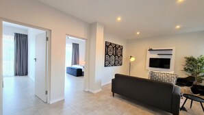 A modern living room with a sofa, a TV, and a floor lamp. There is a door leading to another room and a plant in the corner. - MINT Apartments Greenside (Randburg)