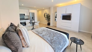 A modern bedroom with a large bed, a small dining table, a TV, and a kitchenette in the background. - MINT Apartments Greenside (Randburg)