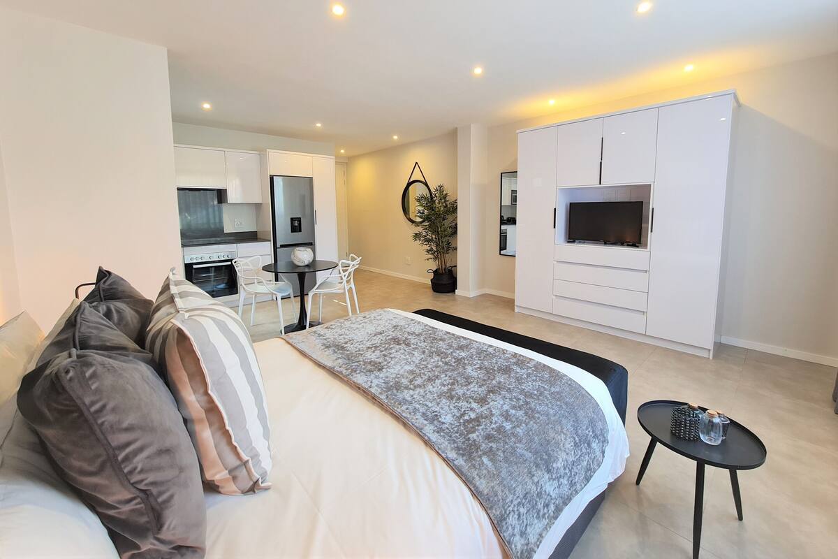 Een moderne slaapkamer met een groot bed, een kleine eettafel, een televisie en een kitchenette op de achtergrond.