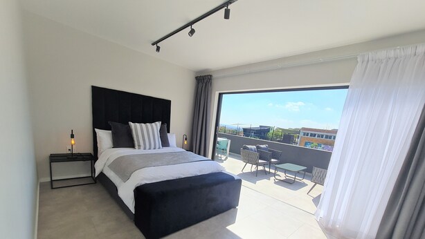 A modern bedroom with a large bed, a bedside table with a lamp, and a balcony with outdoor furniture. - MINT Apartments Greenside (Randburg)
