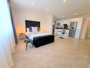 Quarto de hotel moderno com uma cama grande, cabeceira preta, mesa redonda pequena com luminária, banqueta preta e uma cozinha compacta ao fundo.