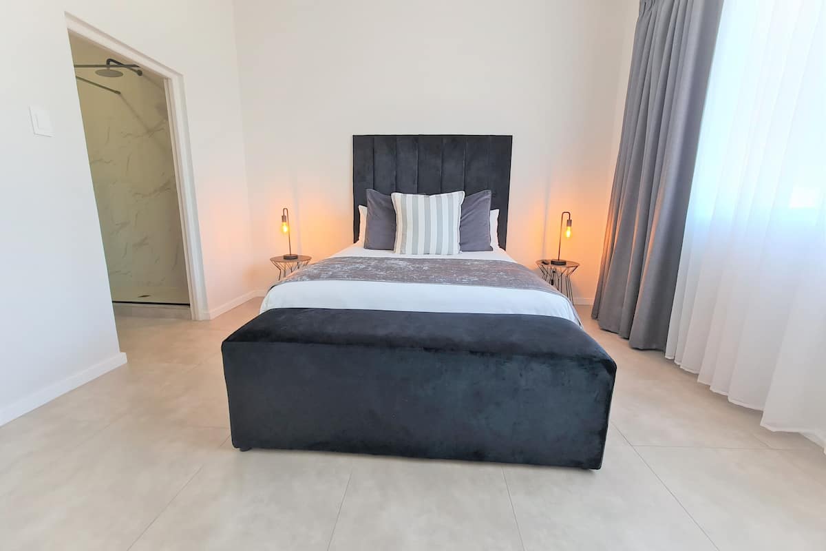 Een moderne slaapkamer met een groot bed, twee nachtkastjes met lampen, een donkere hoofdbord en uitzicht op een badkamer door een open deur.