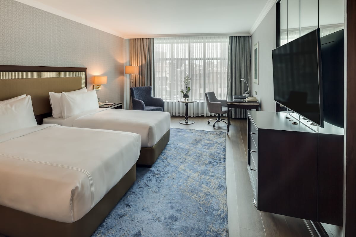 Habitación de hotel con dos camas, un escritorio, un televisor y vistas a la ciudad a través de amplias ventanas.