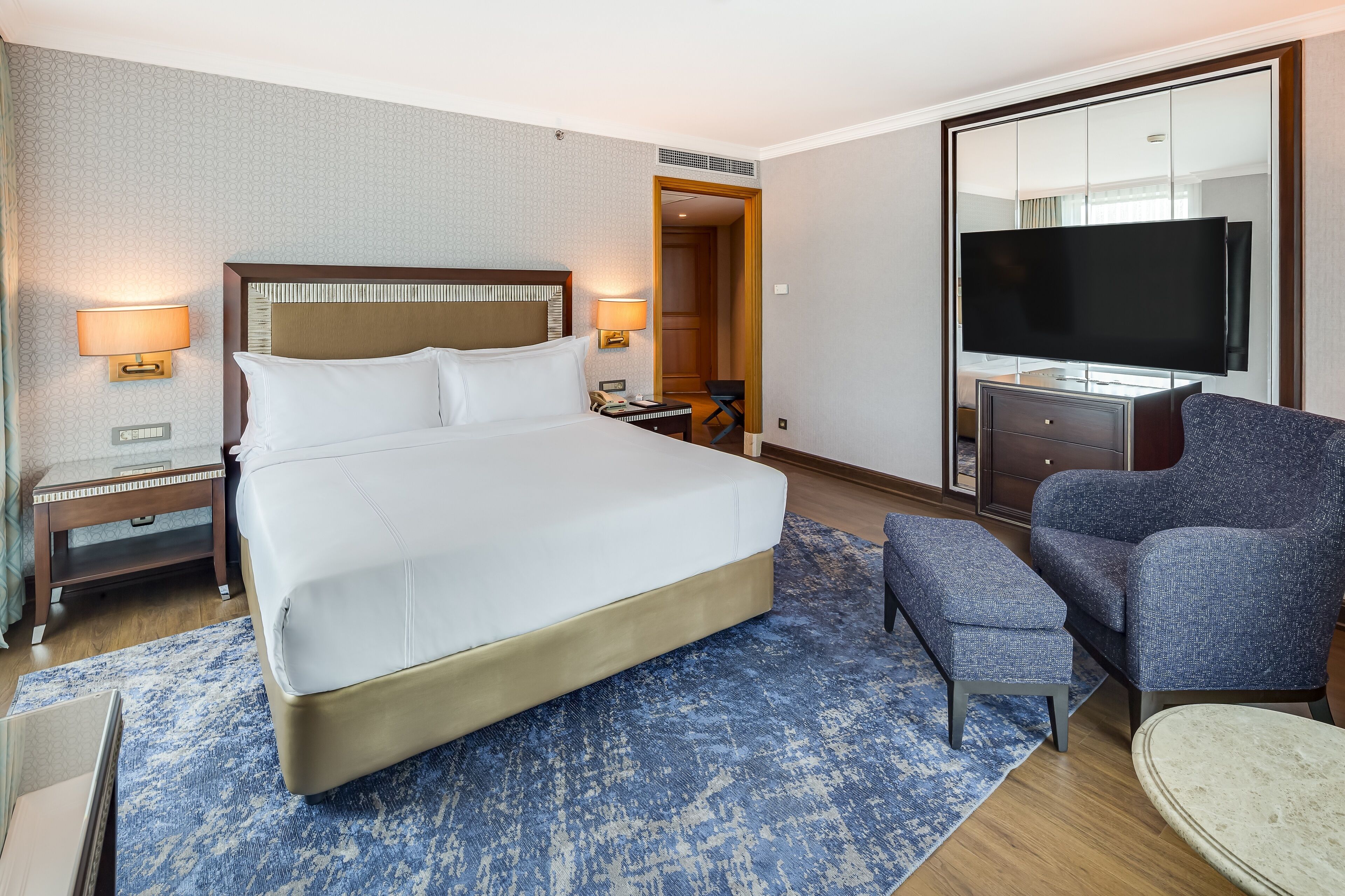 A hotel room with a large bed, a desk with a lamp, a television, and a chair.