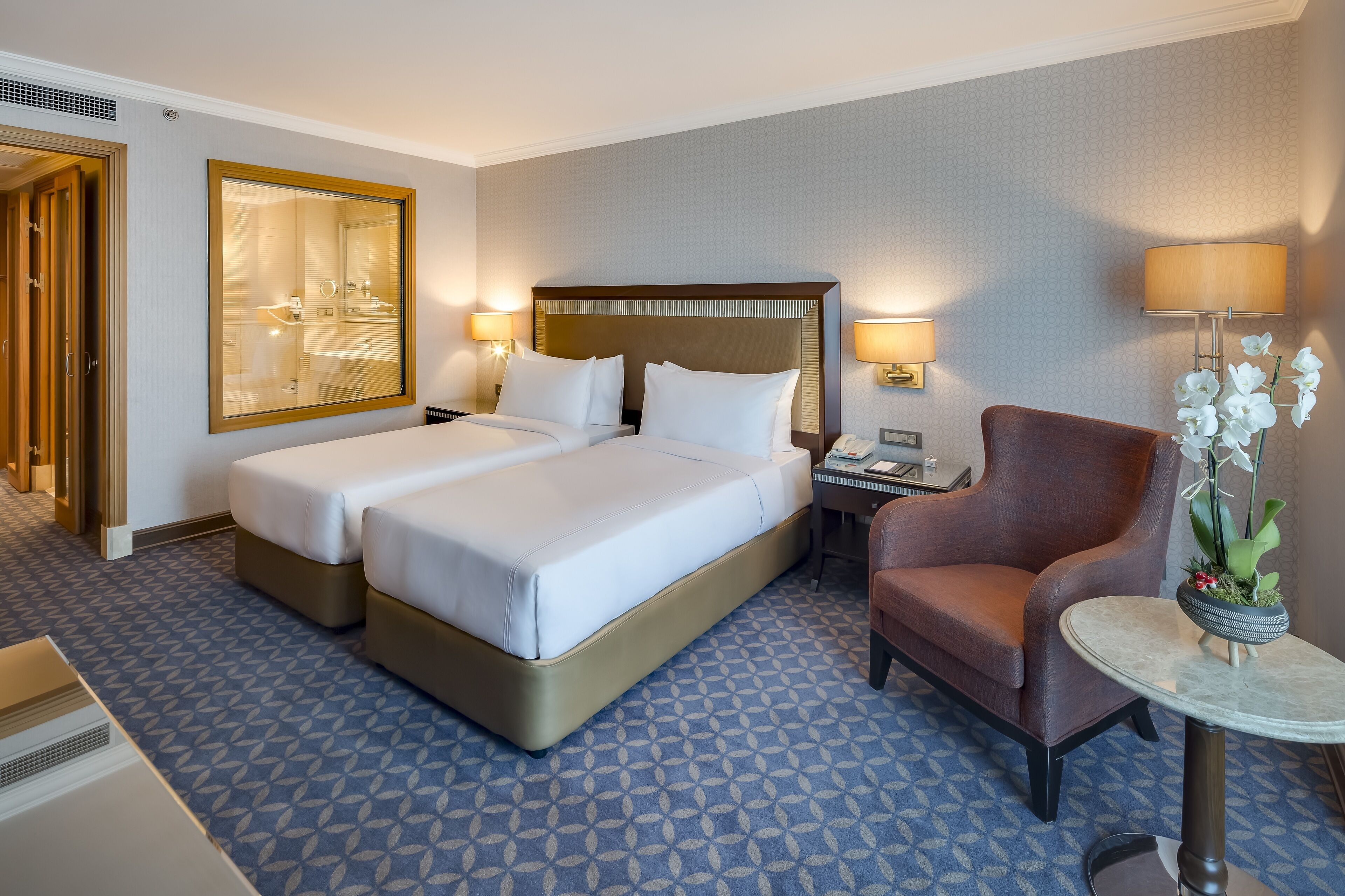 A hotel room with a double bed, a desk with a lamp, a chair, and a small table with a plant.
