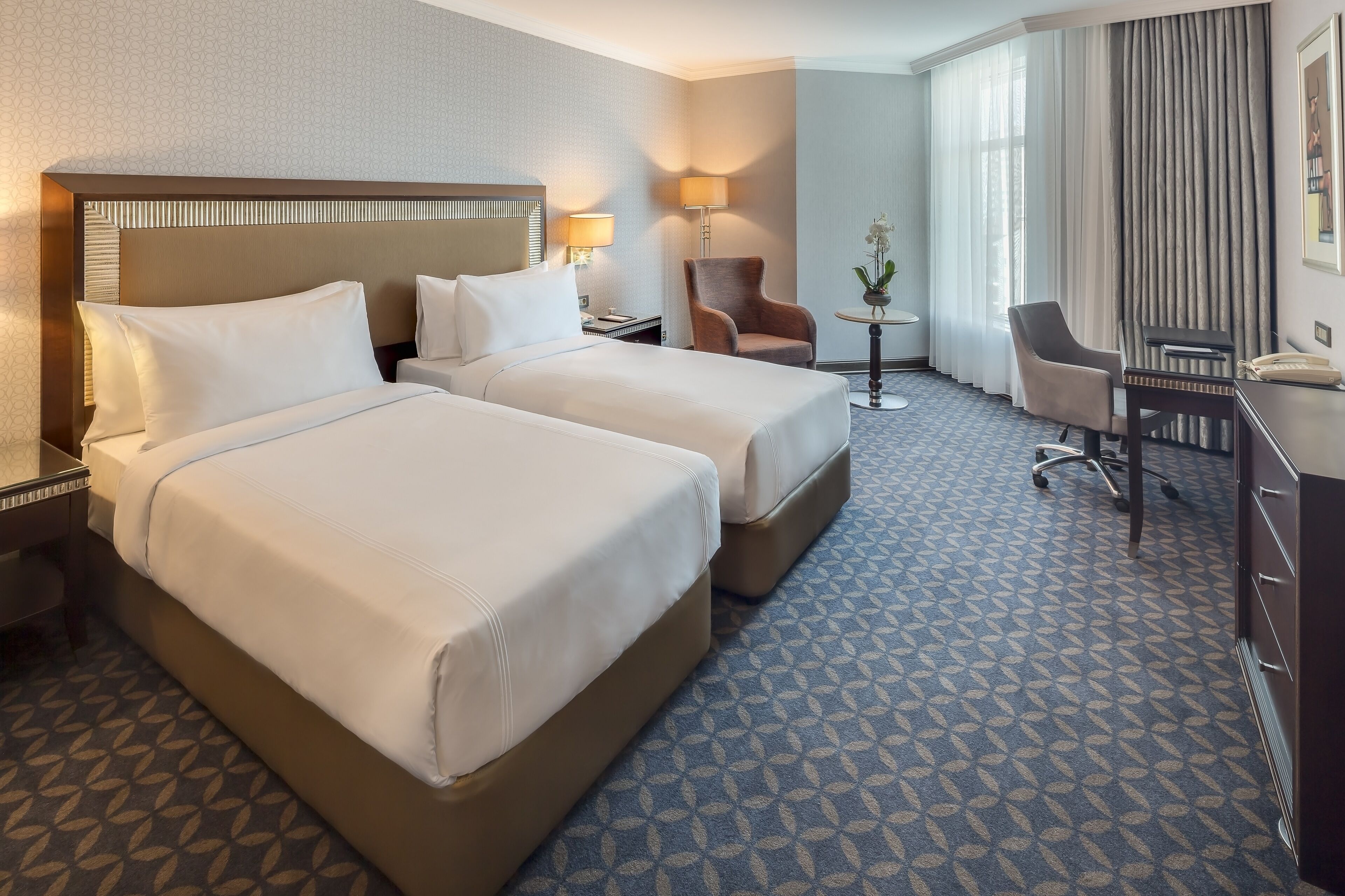 A hotel room with two beds, a desk, a chair, and a small table with a lamp.