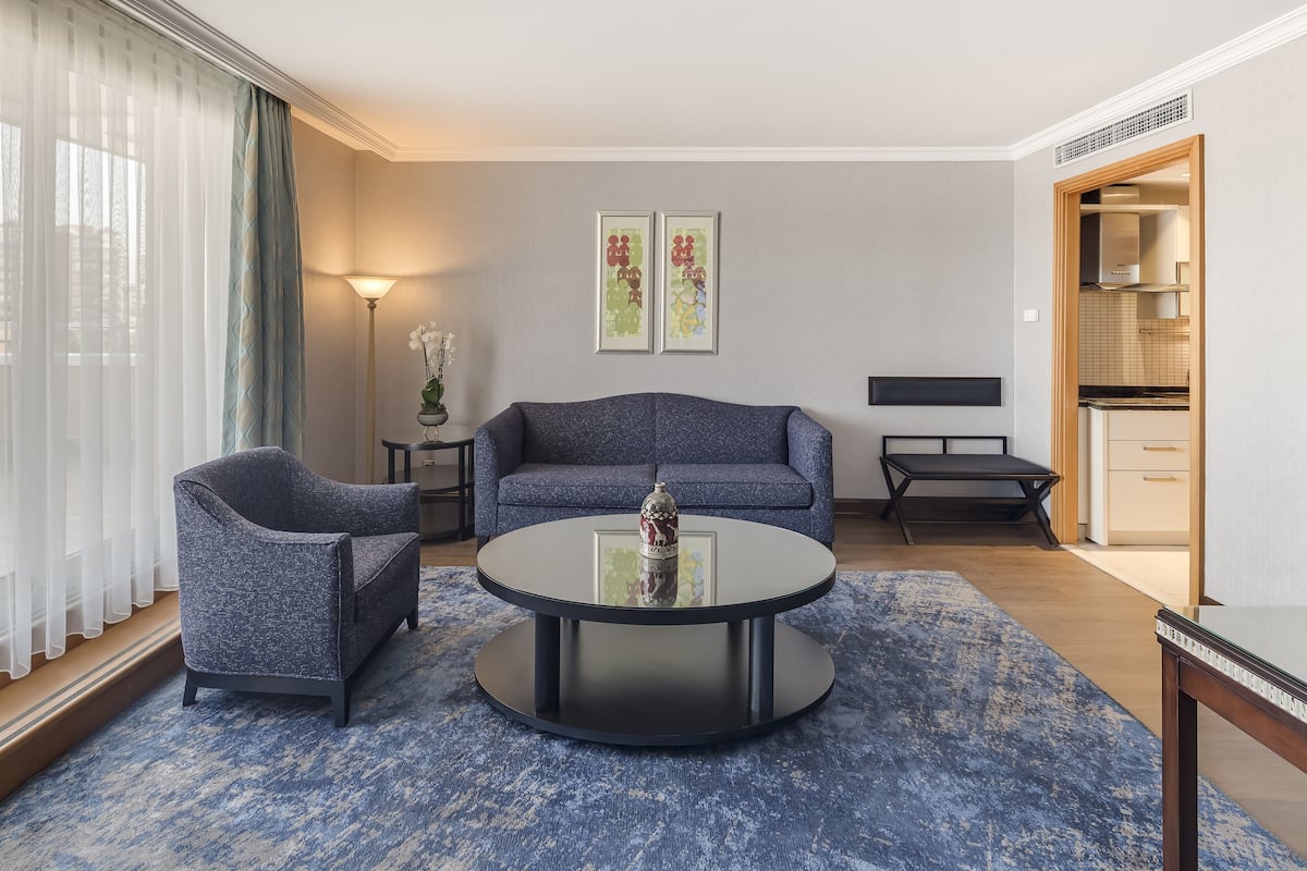 Una sala moderna con una mesa redonda, dos sillones y un sofá. Hay dos cuadros enmarcados en la pared, una lámpara de pie y vistas a la cocina.
