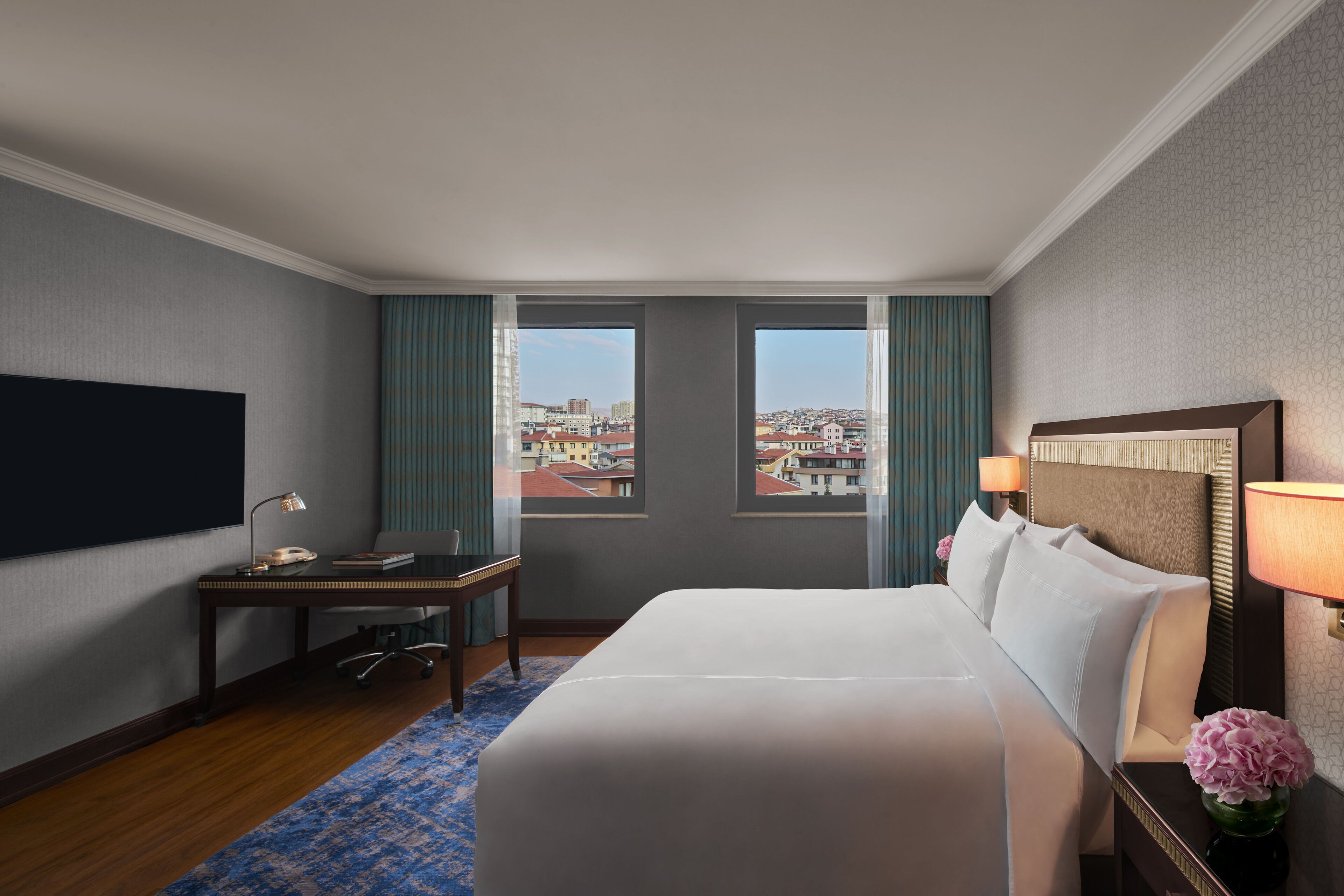 A hotel room with a large bed, a desk with a lamp, a TV, and a view of the city through two windows.