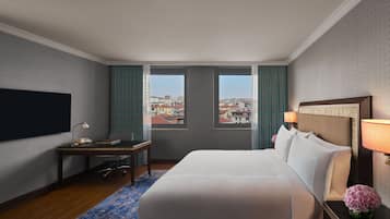 Ein Hotelzimmer mit einem großen Bett, einem Schreibtisch mit Lampe, einem Fernseher und Blick auf die Stadt durch zwei Fenster.