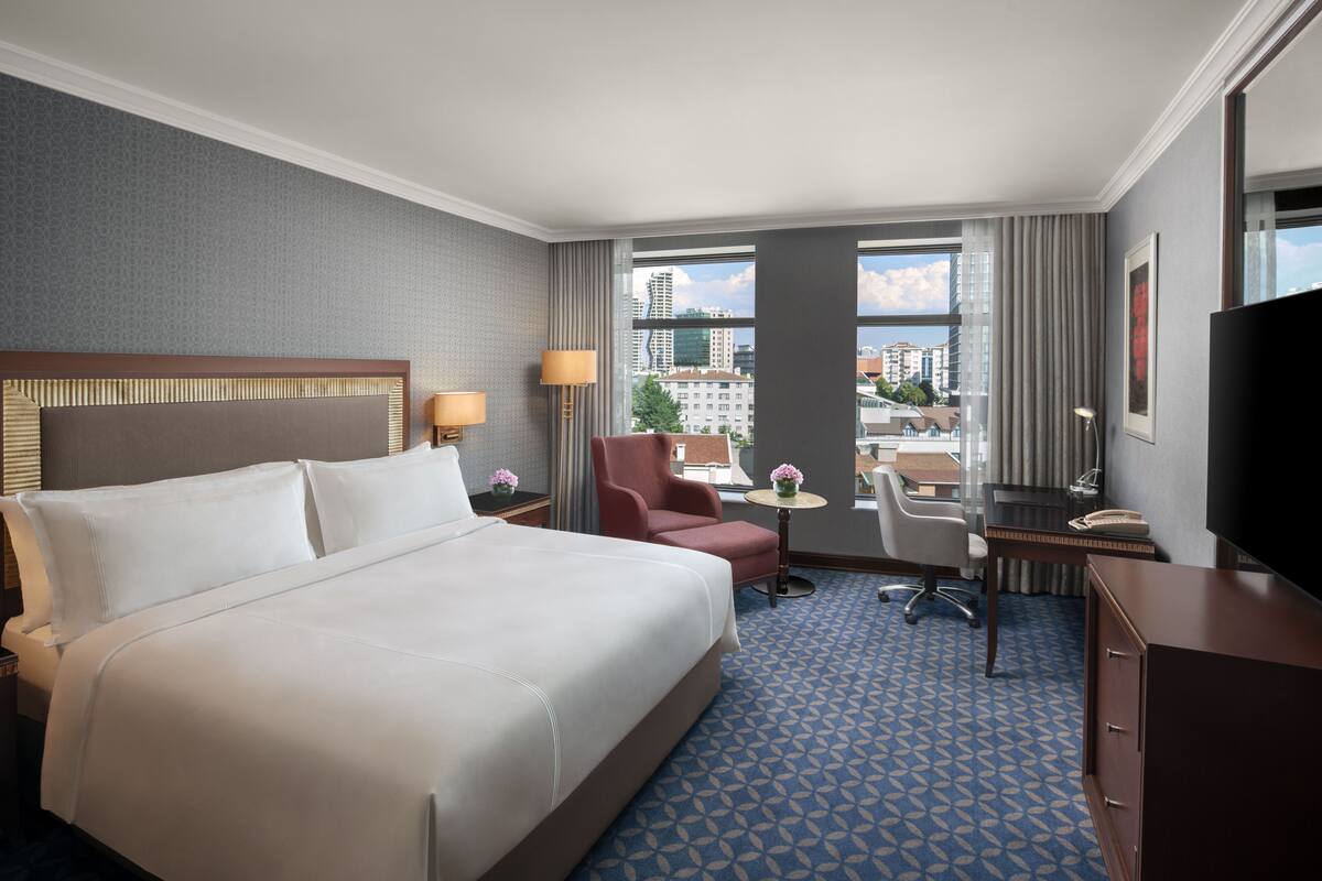 Habitación de hotel con una cama grande, un escritorio, una silla, un televisor y vistas a la ciudad desde las ventanas.