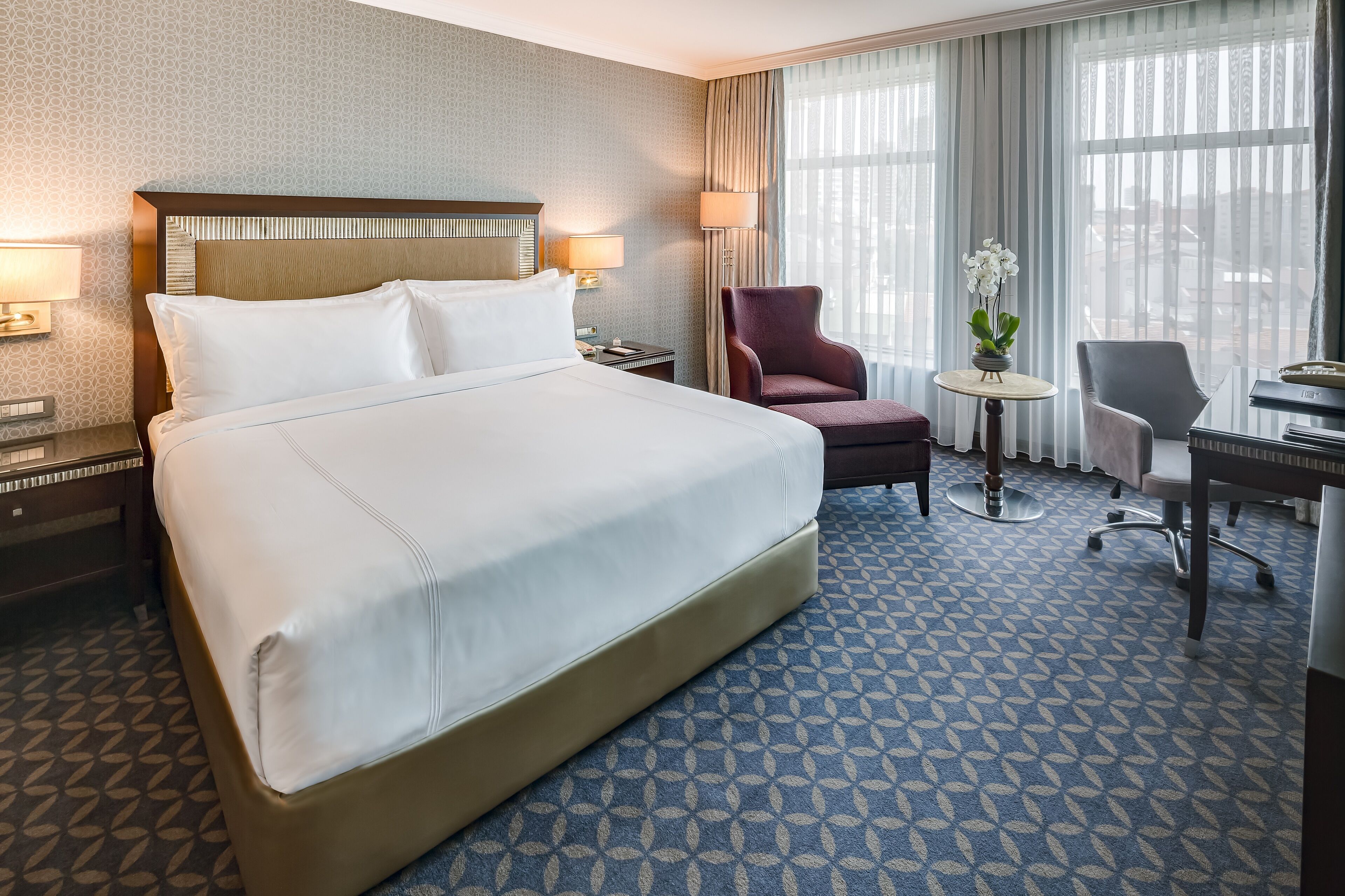 A hotel room with a large bed, two bedside tables with lamps, a desk with a chair, a small round table with a vase, and a view of the city through large windows.