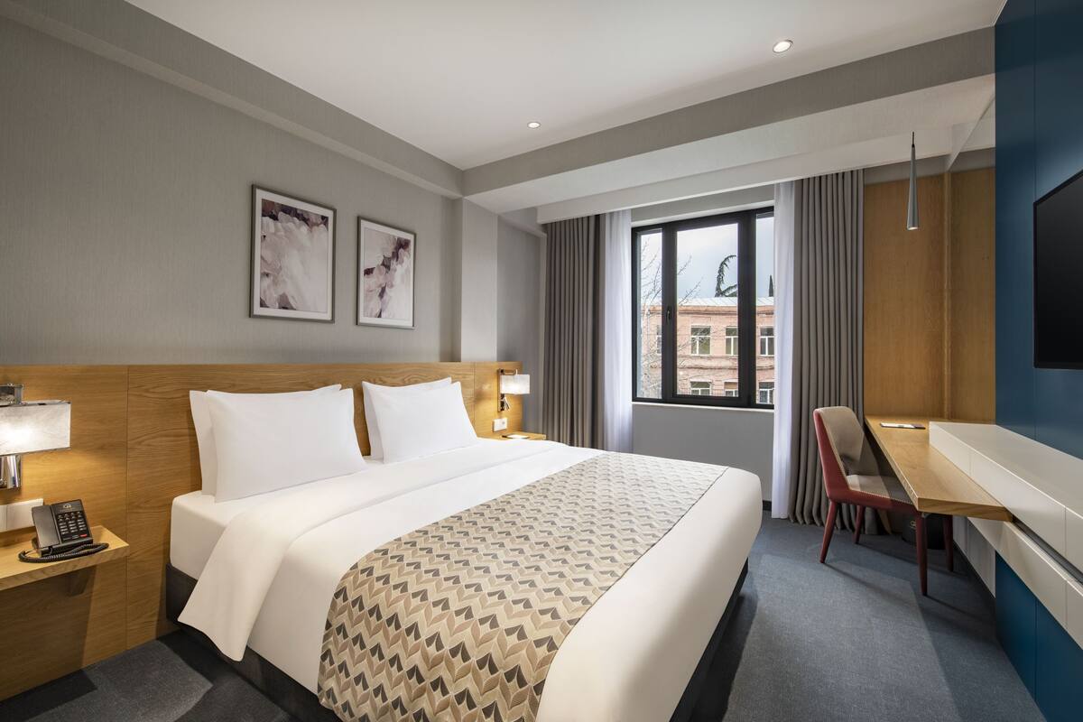 A hotel room with a large bed, a desk with a chair, a telephone, and a window with curtains.