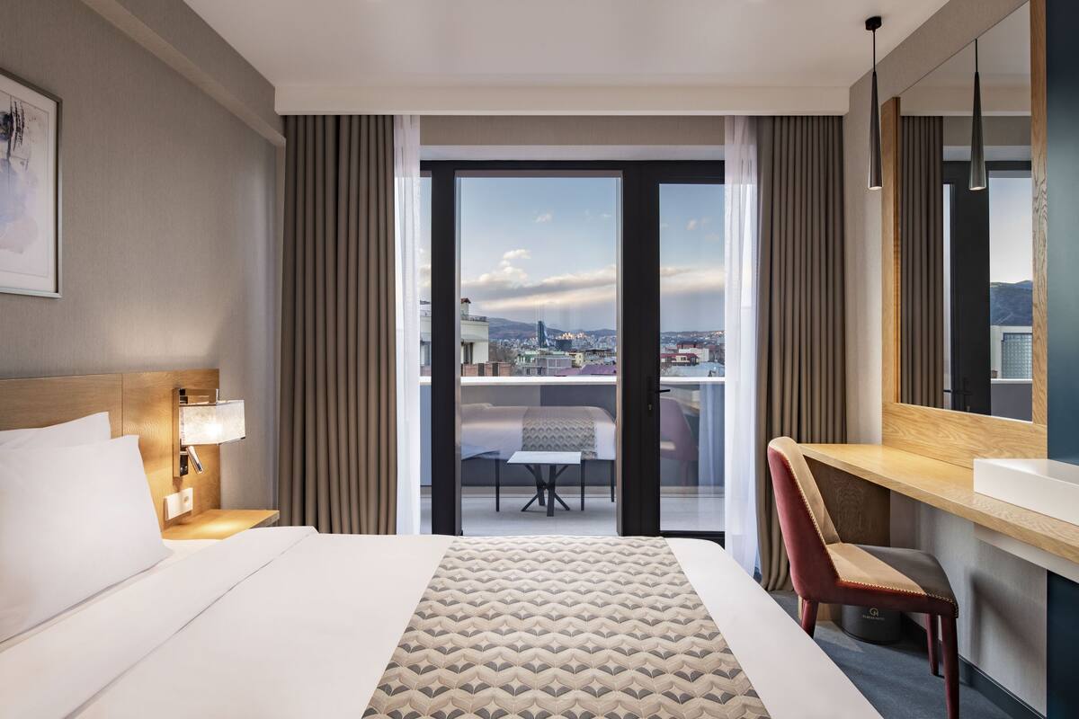 A hotel room with a bed, a desk, a chair, a large window, and a view of the city.