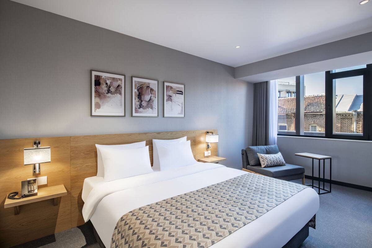 A hotel room with a large bed, bedside tables, a desk, and a view of buildings through the window.