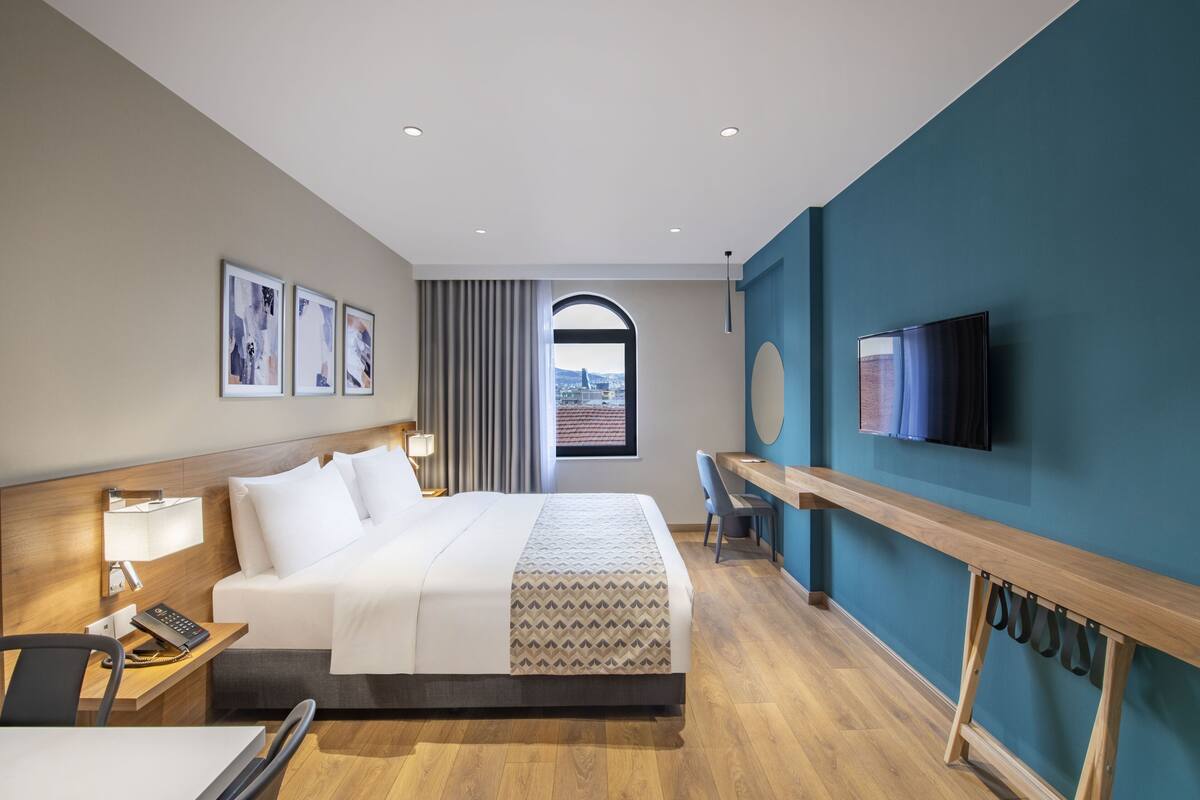 A hotel room with a large bed, a desk with a chair, a TV mounted on the wall, and a window with a view of a cityscape.