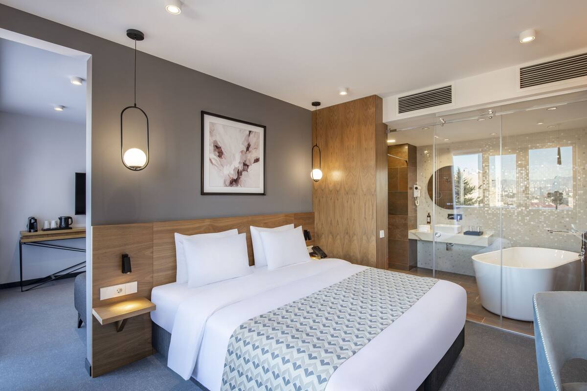 A modern hotel room with a large bed, bedside tables, a desk, a large window, and a bathroom with a bathtub and shower.