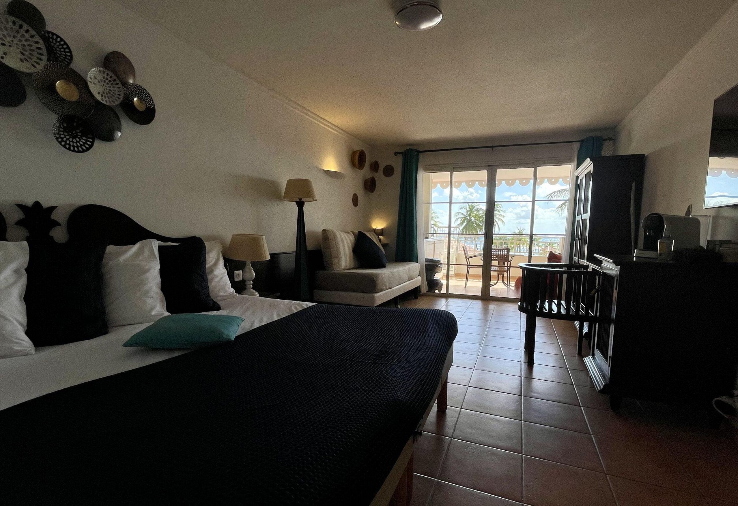 A hotel room with a bed, a desk, a chair, a TV, and a balcony with a view of palm trees.