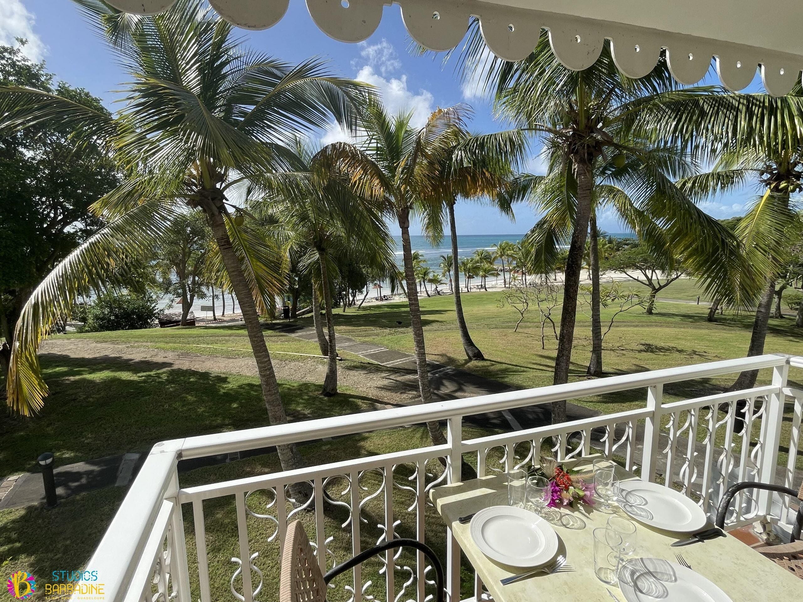 A balcony with a table set for two, overlooking a beach with palm trees and the ocean.