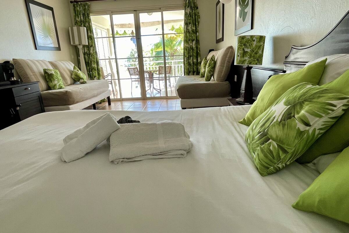 Ein Hotelzimmer mit Bett, zwei Sesseln, einem Tisch und einem Balkon mit Blick auf Grünflächen.