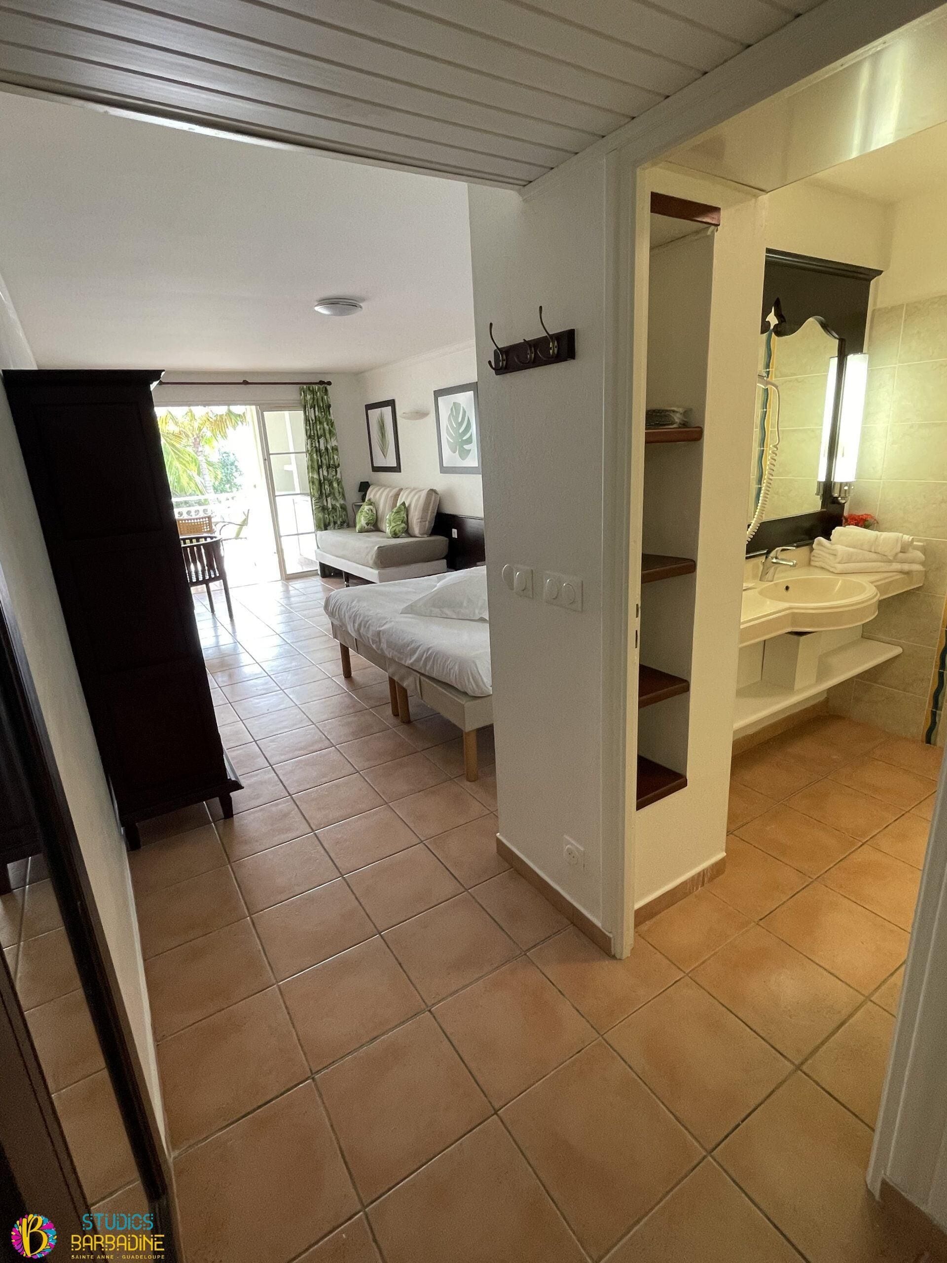 Une chambre d’hôtel comprenant un lit, un petit coin salon avec un canapé et des coussins, une salle de bain avec un lavabo et un miroir, et une vue sur l’extérieur.