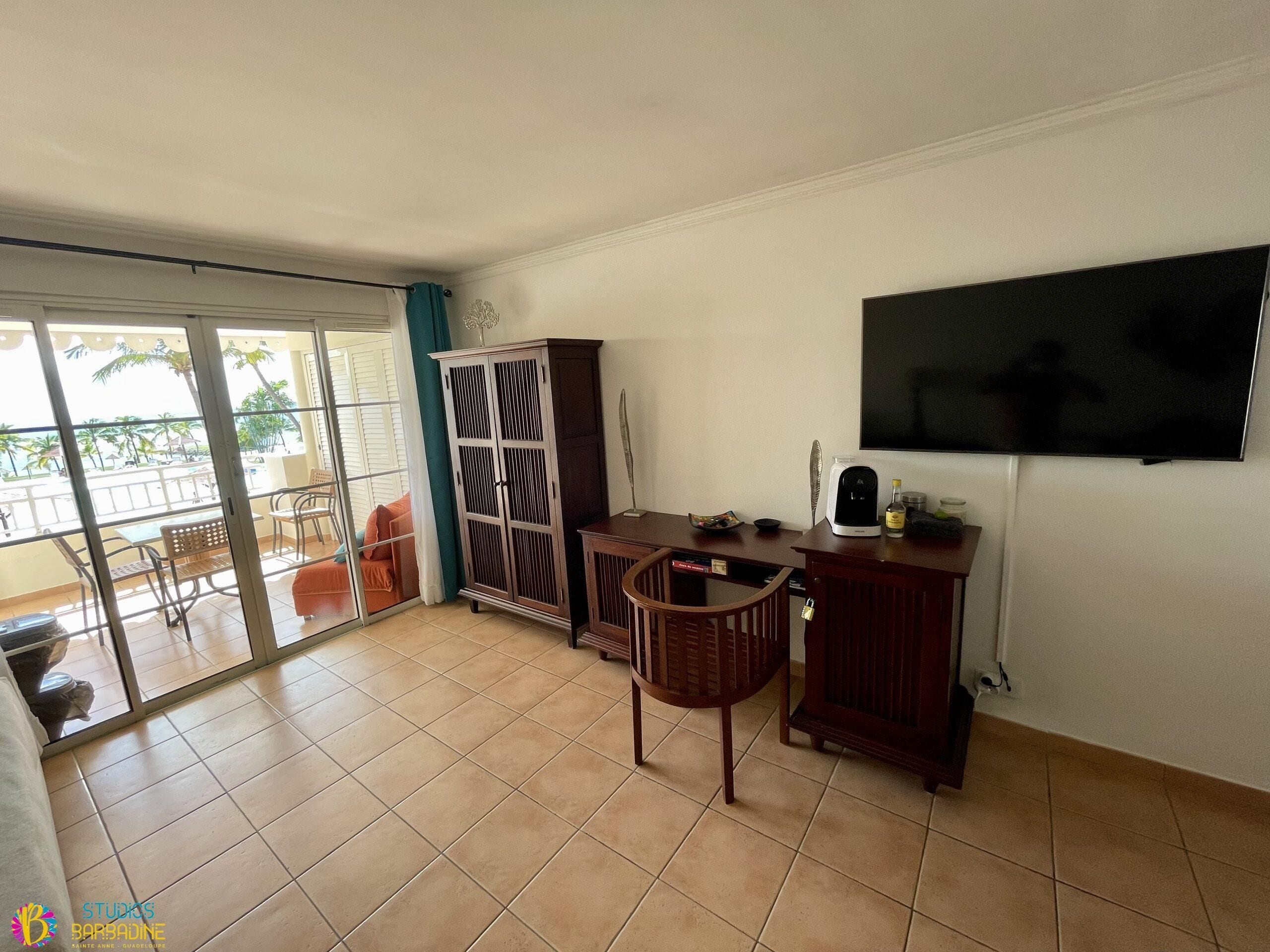A room with a sliding glass door leading to a balcony, a TV mounted on the wall, a wooden cabinet, and a small table with chairs.