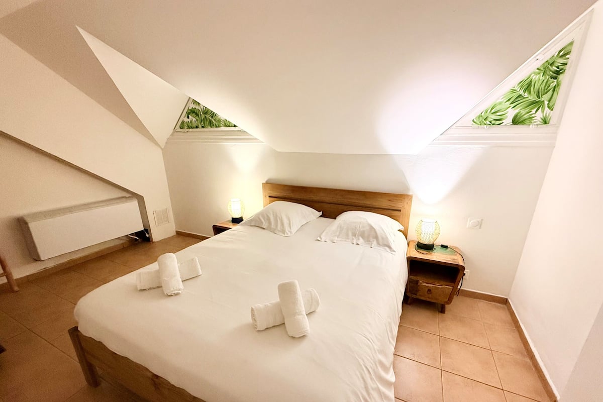 Ein Schlafzimmer mit einem Bett, zwei Nachttischen, einem Heizkörper und einem Fenster mit Blick auf Pflanzen.