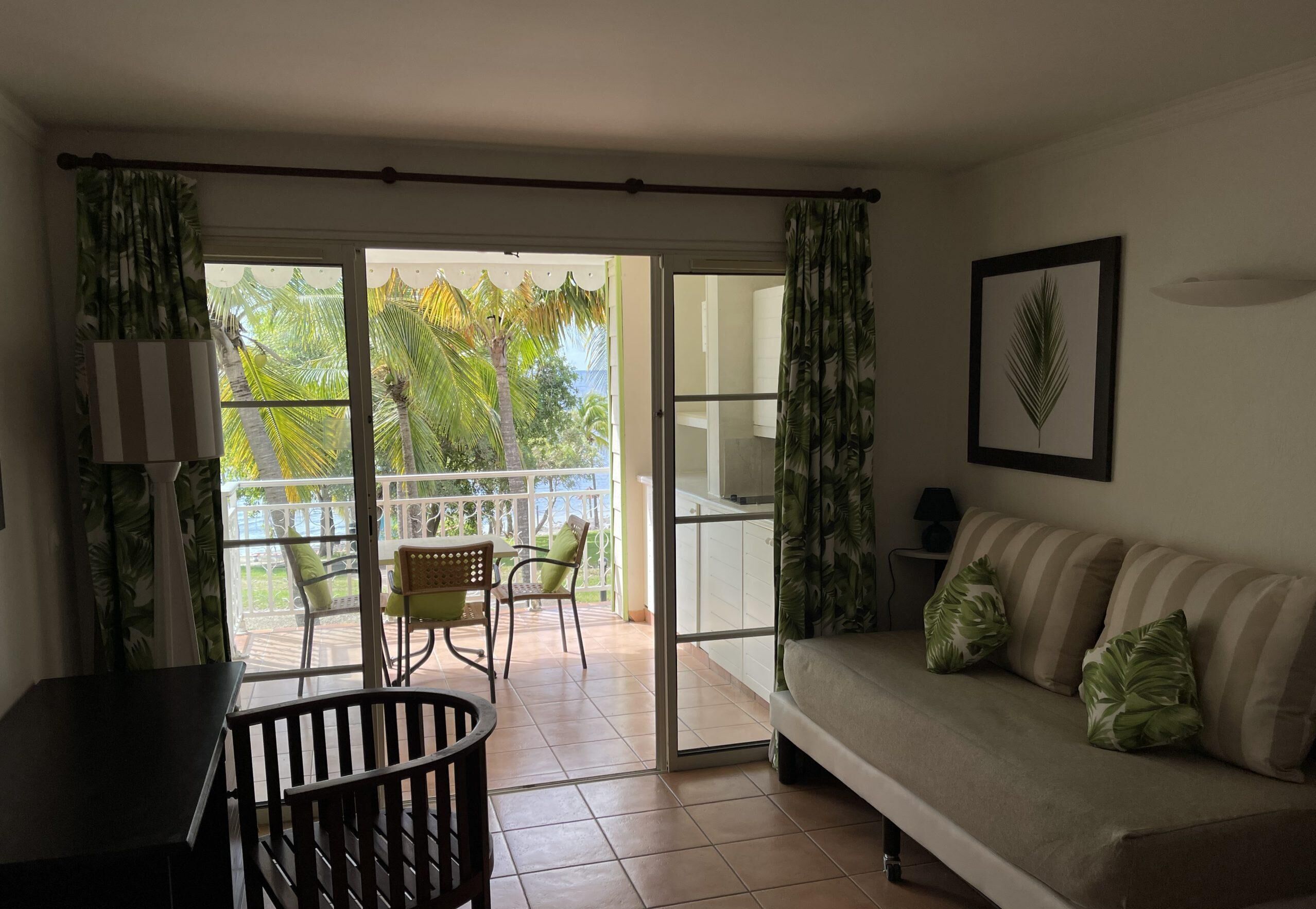 A living room with a sofa, a dining table, and chairs. There is a balcony with a view of palm trees and a white railing.