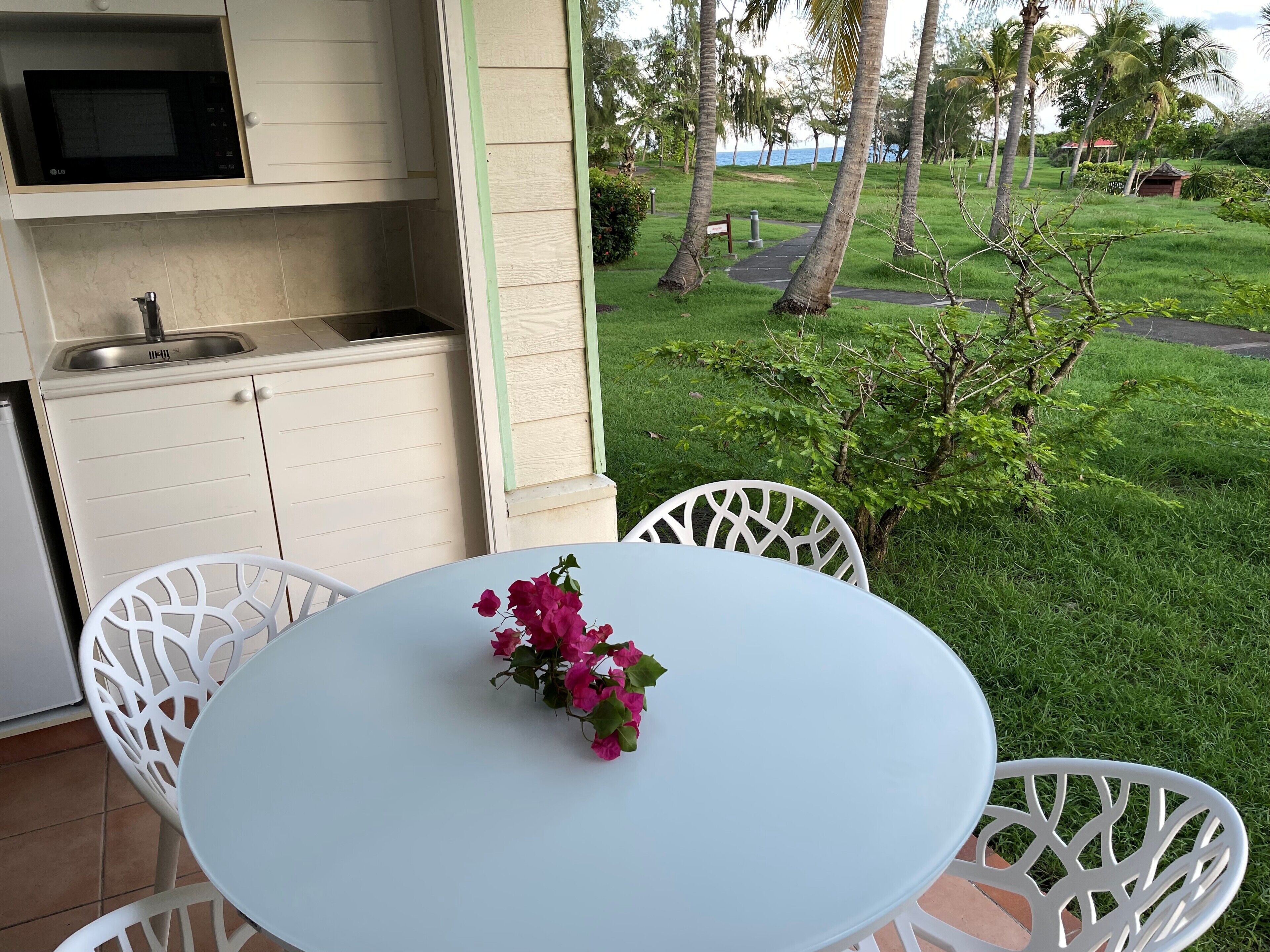 A kitchen with a microwave, sink, and cabinets, a white table with chairs, and a view of a grassy area with trees and a path.