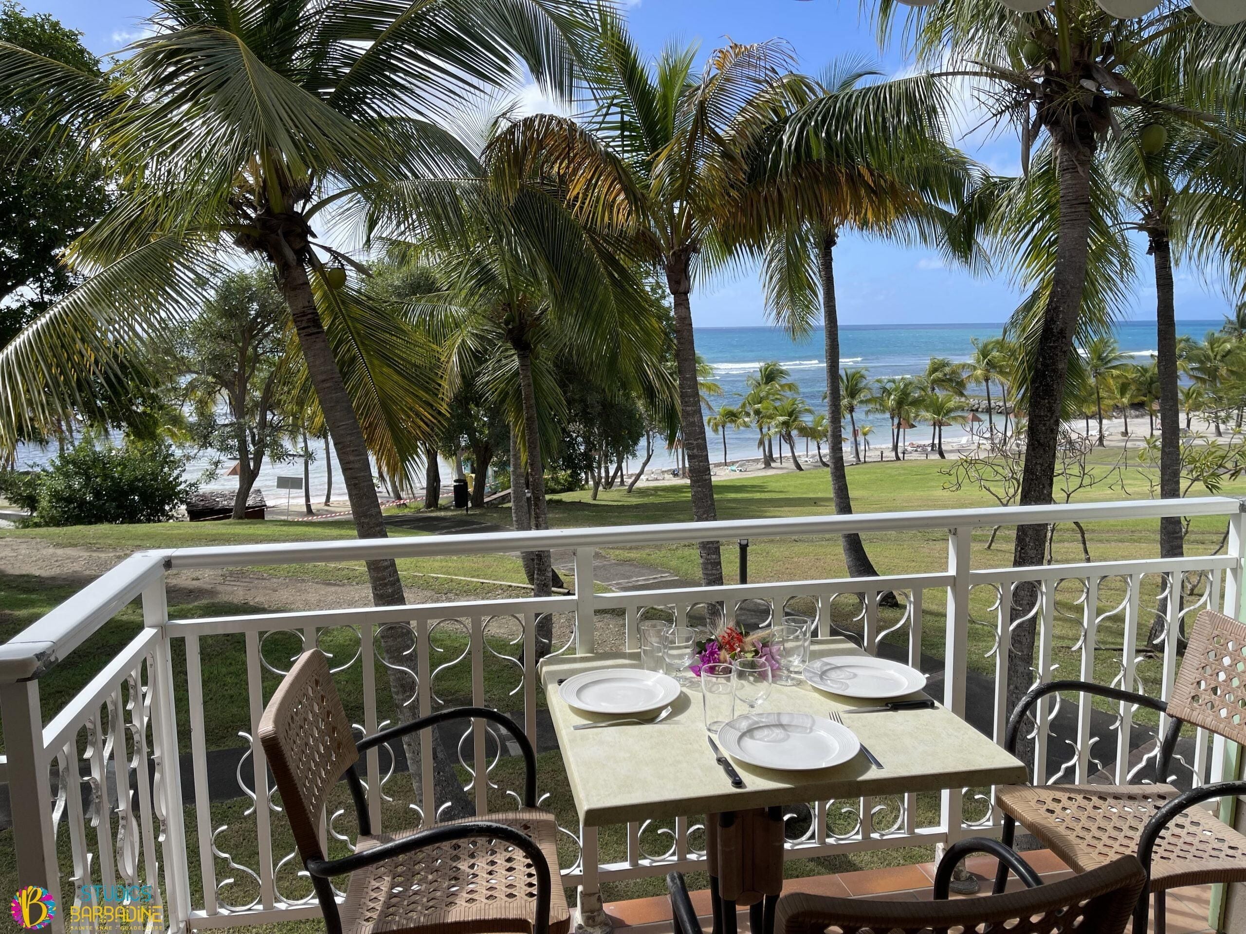 A balcony with a table set for two, overlooking a beach with palm trees and the ocean.