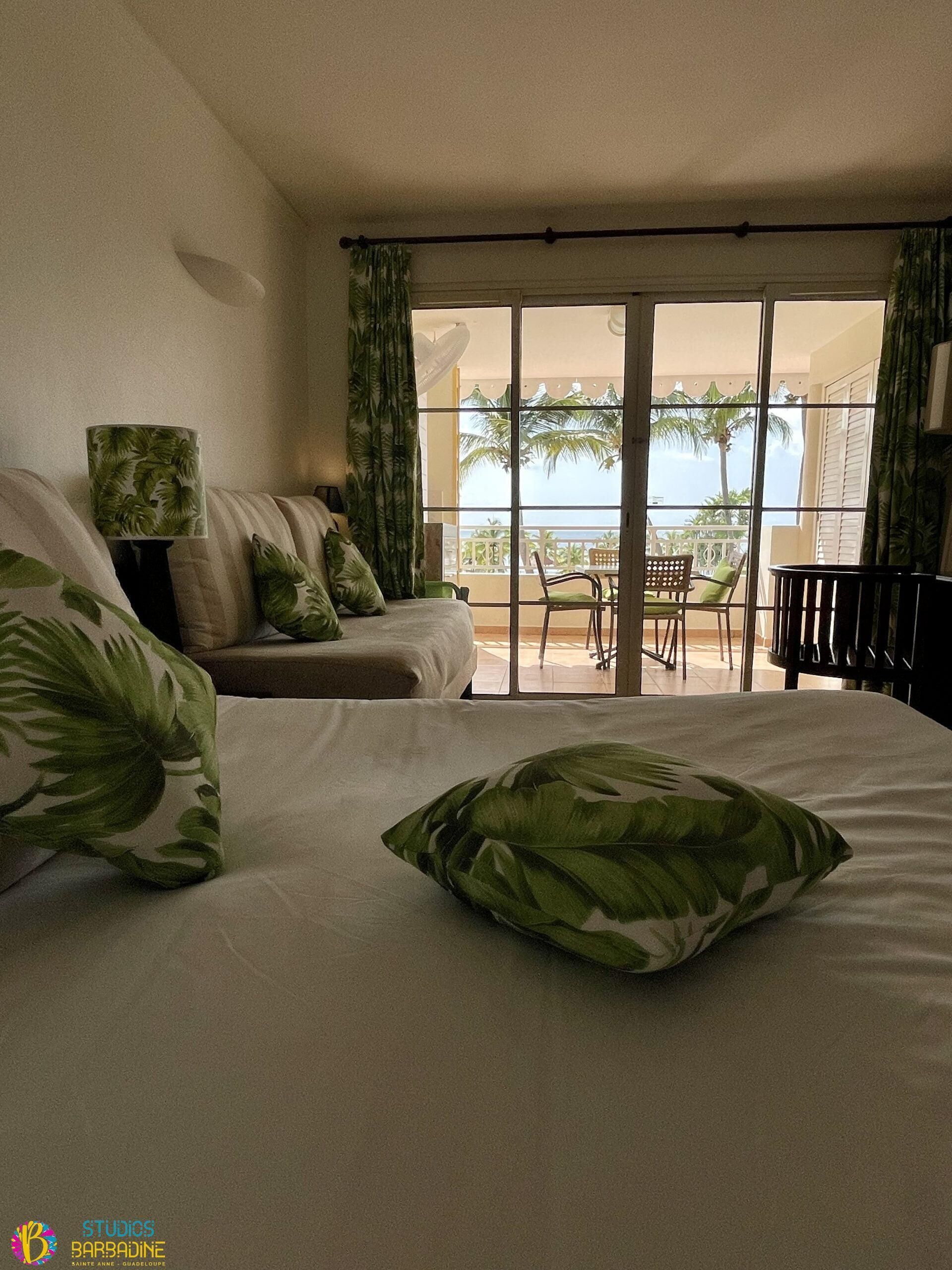 Habitación de hotel con cama, sofá, mesa y sillón. Tiene un balcón con vistas a palmeras y al mar.