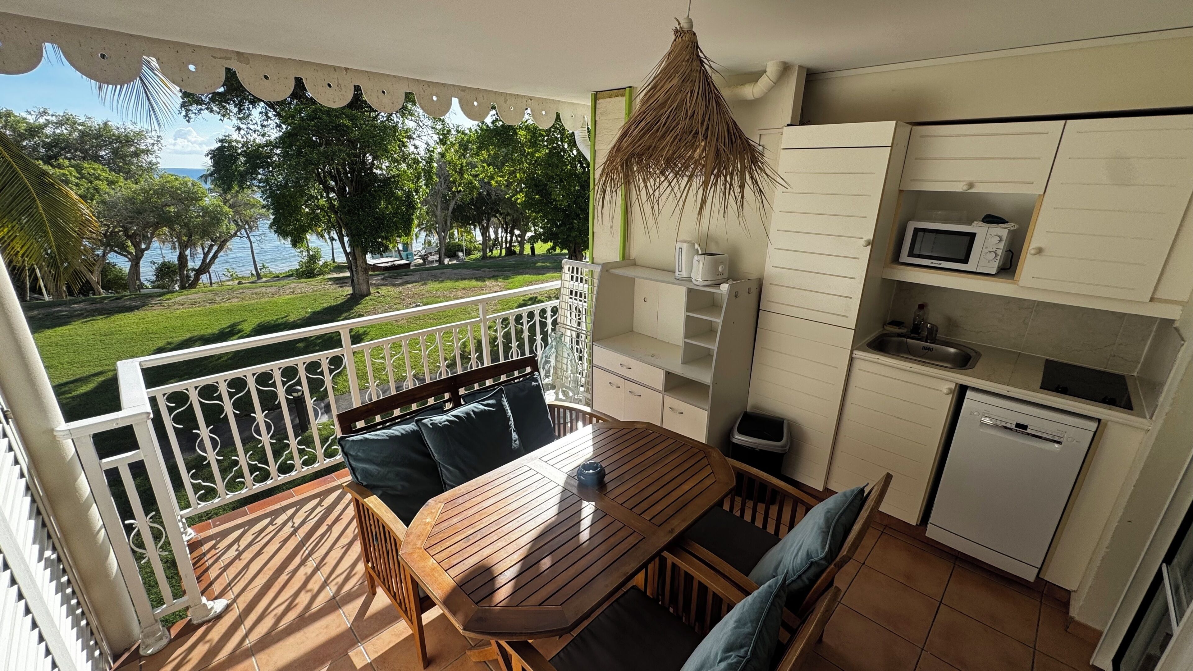 A balcony with a wooden table and chairs, a small kitchen area with a microwave, and a view of a grassy area with trees.