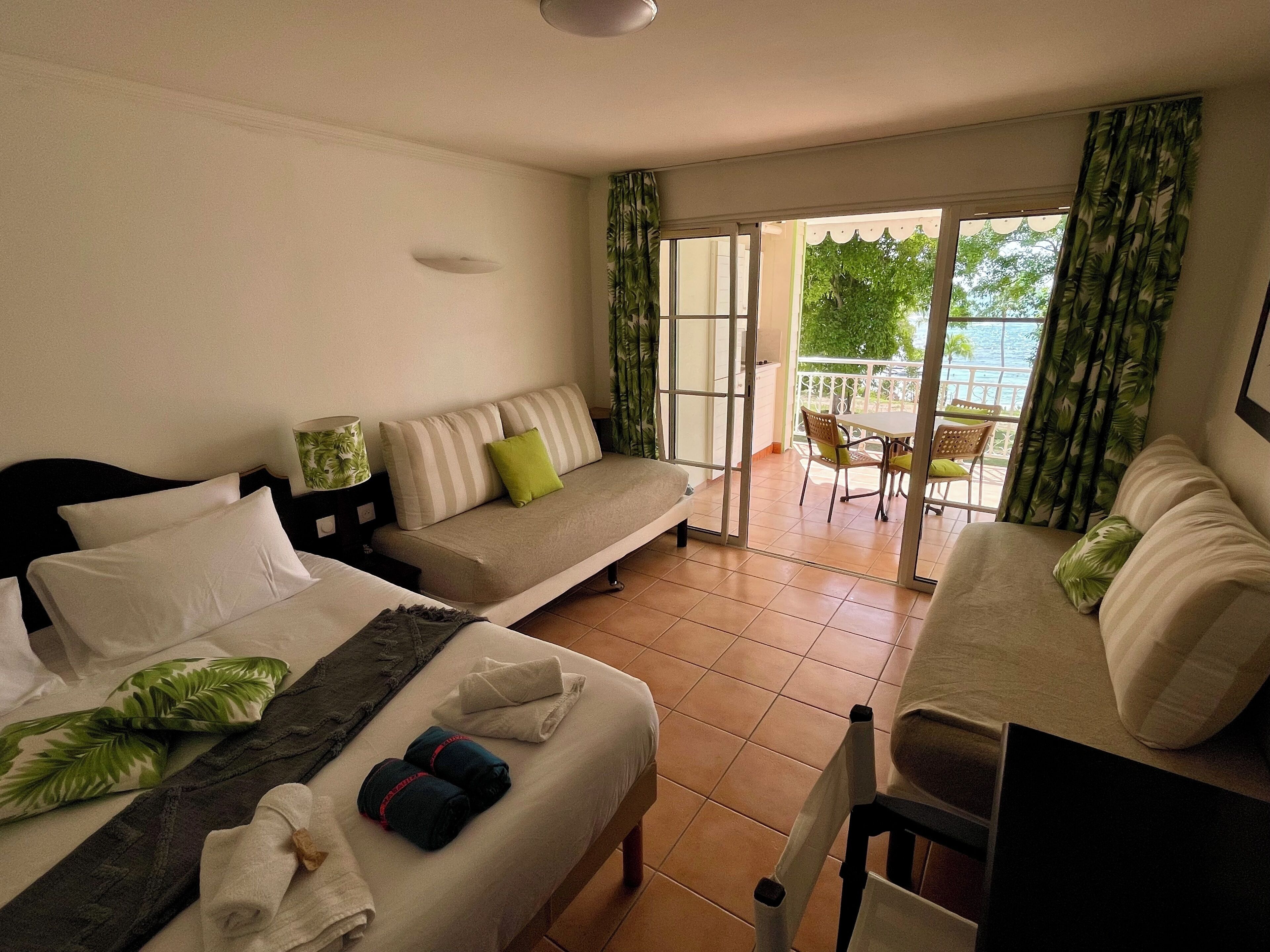 A hotel room with a bed, a sofa, a dining table, and a balcony with a view of the outdoors.
