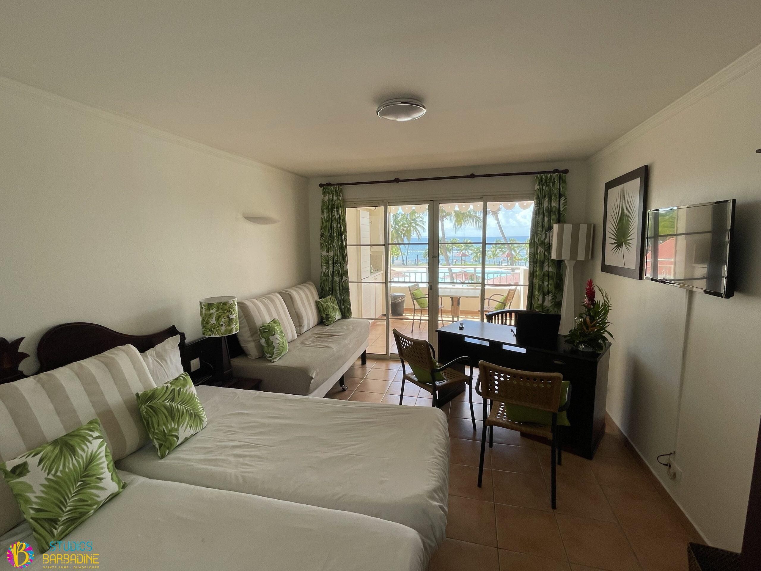 Habitación con sofá, cama, escritorio y televisor. Se ve una ventana con cortinas y vista a palmeras.