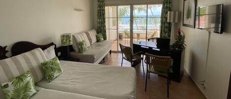 A room with a sofa, a bed, a desk, and a TV. There is a window with curtains and a view of palm trees.