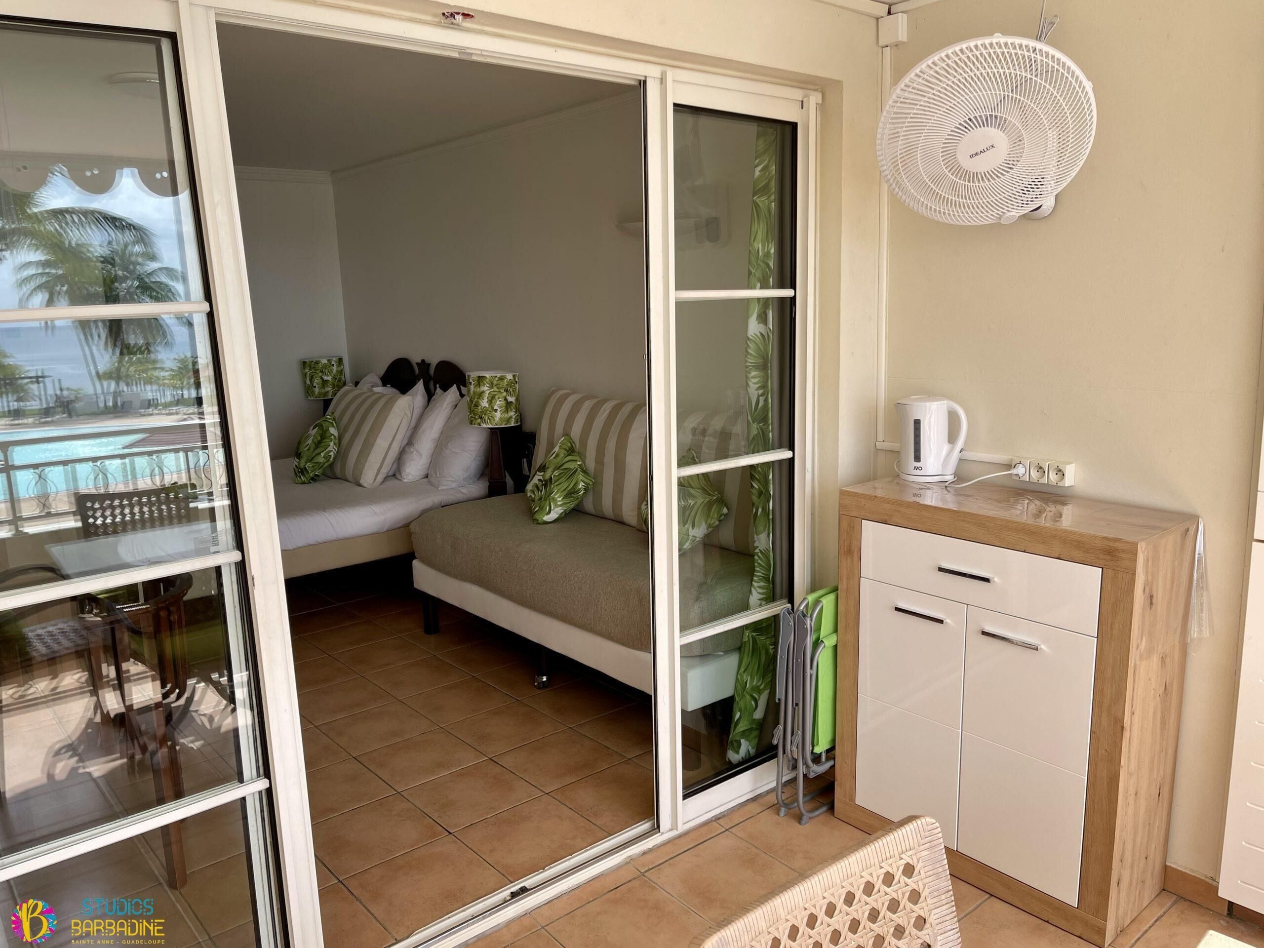 A room with a bed, a small desk, a fan, and a view of the outside.