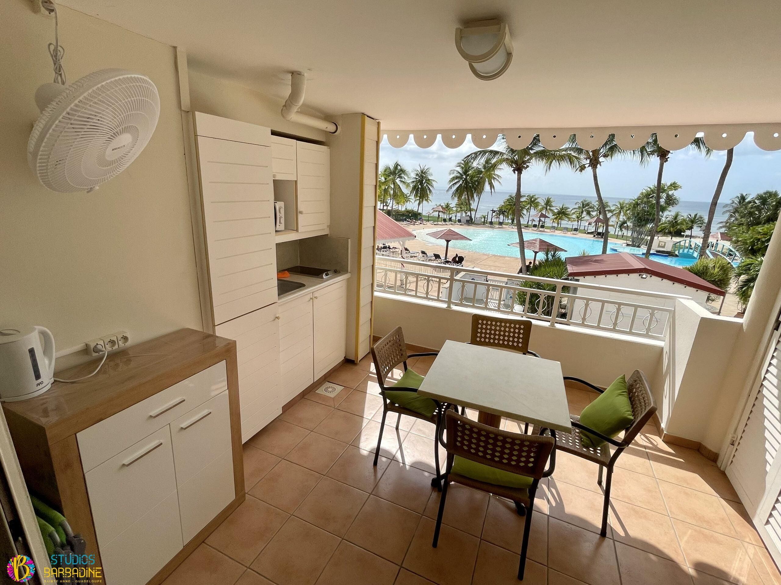 A small balcony with a view of a pool and palm trees, a kitchenette with a fan, and a table with chairs.
