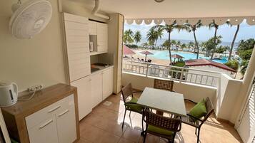 A small balcony with a view of a pool and palm trees, a kitchenette with a fan, and a table with chairs.
