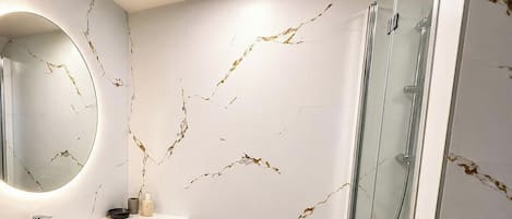 Ein Badezimmer mit weißer Marmorwand, einem runden Spiegel, einem Waschbecken und einer Duschkabine aus Glas.