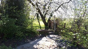 Ein gepflasterter Weg führt zu einer Holzhütte, umgeben von üppiger Vegetation und blühenden Bäumen.