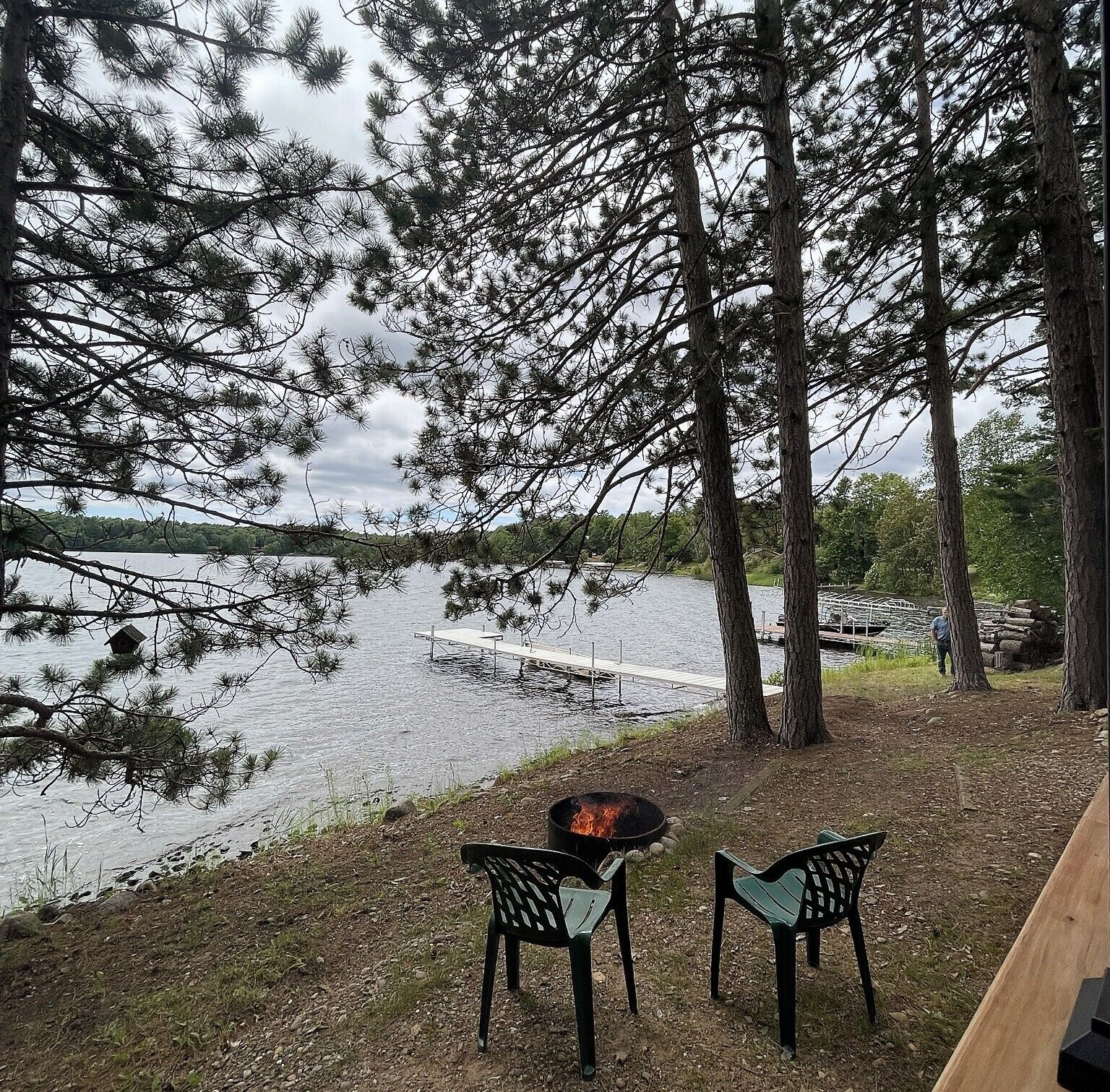 Ein Sitzbereich am Seeufer mit Feuerstelle und Stühlen, umgeben von Bäumen und einem Steg, der ins Wasser führt.
