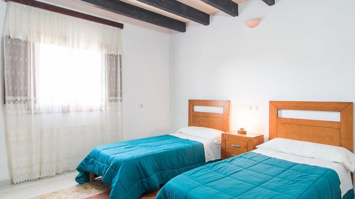 A room with two single beds, wooden headboards, a nightstand with a lamp, and a window with sheer curtains.