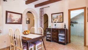 A dining area with a wooden table, chairs, and a fruit bowl. There are two paintings on the walls, a cabinet with glass doors, and a view of a building outside. - Villa with pool very close to Es Trenc Beach (Campos)