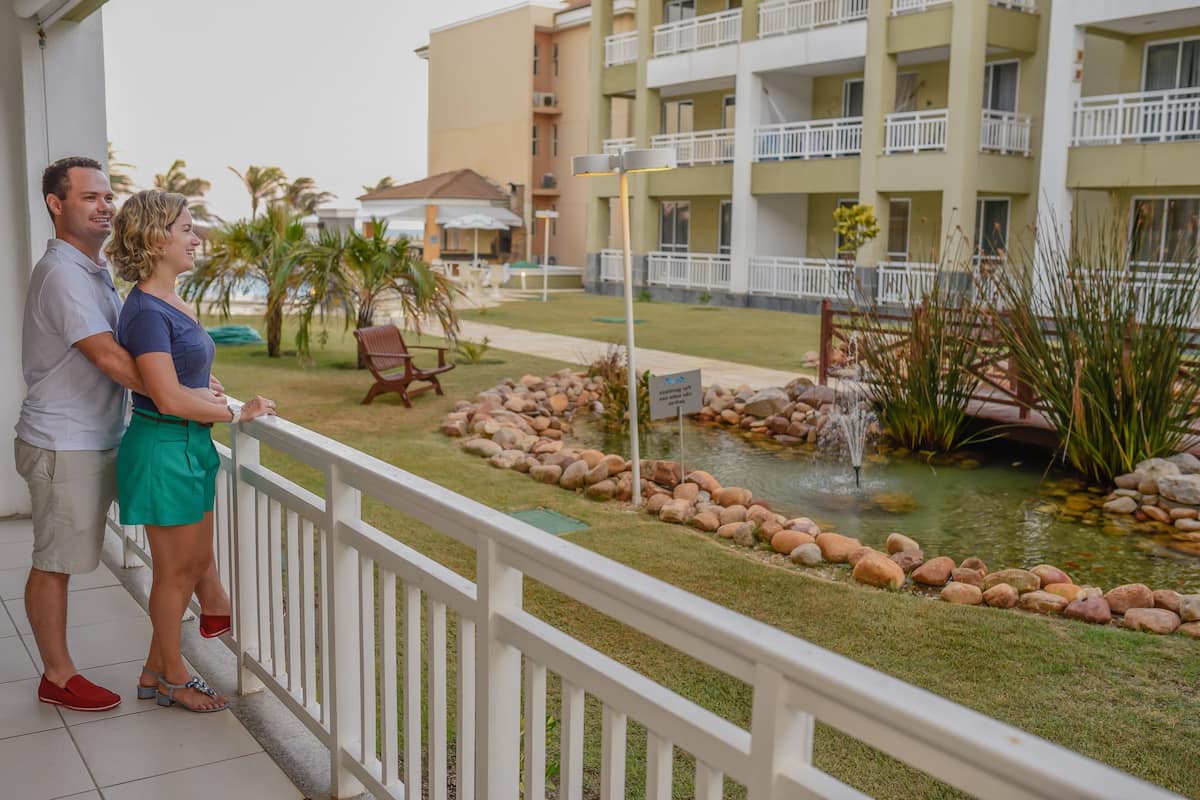 Un couple se tient sur un balcon donnant sur un jardin aménagé avec un étang, le tout entouré d’un immeuble à plusieurs étages doté de balcons et d’espaces de détente en plein air.