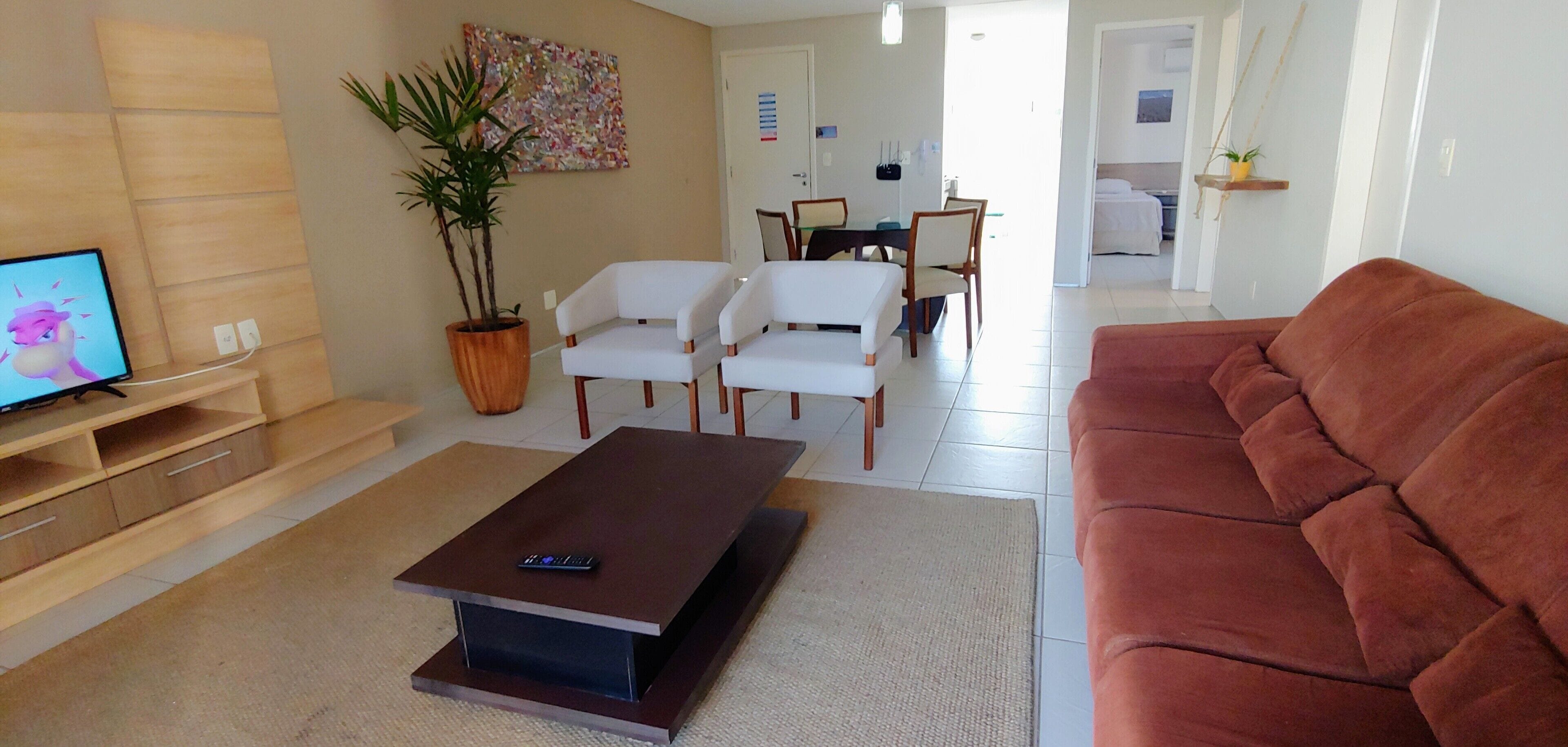 A living room with a television, a brown coffee table, white chairs, a red sofa, a potted plant, and a dining area in the background.
