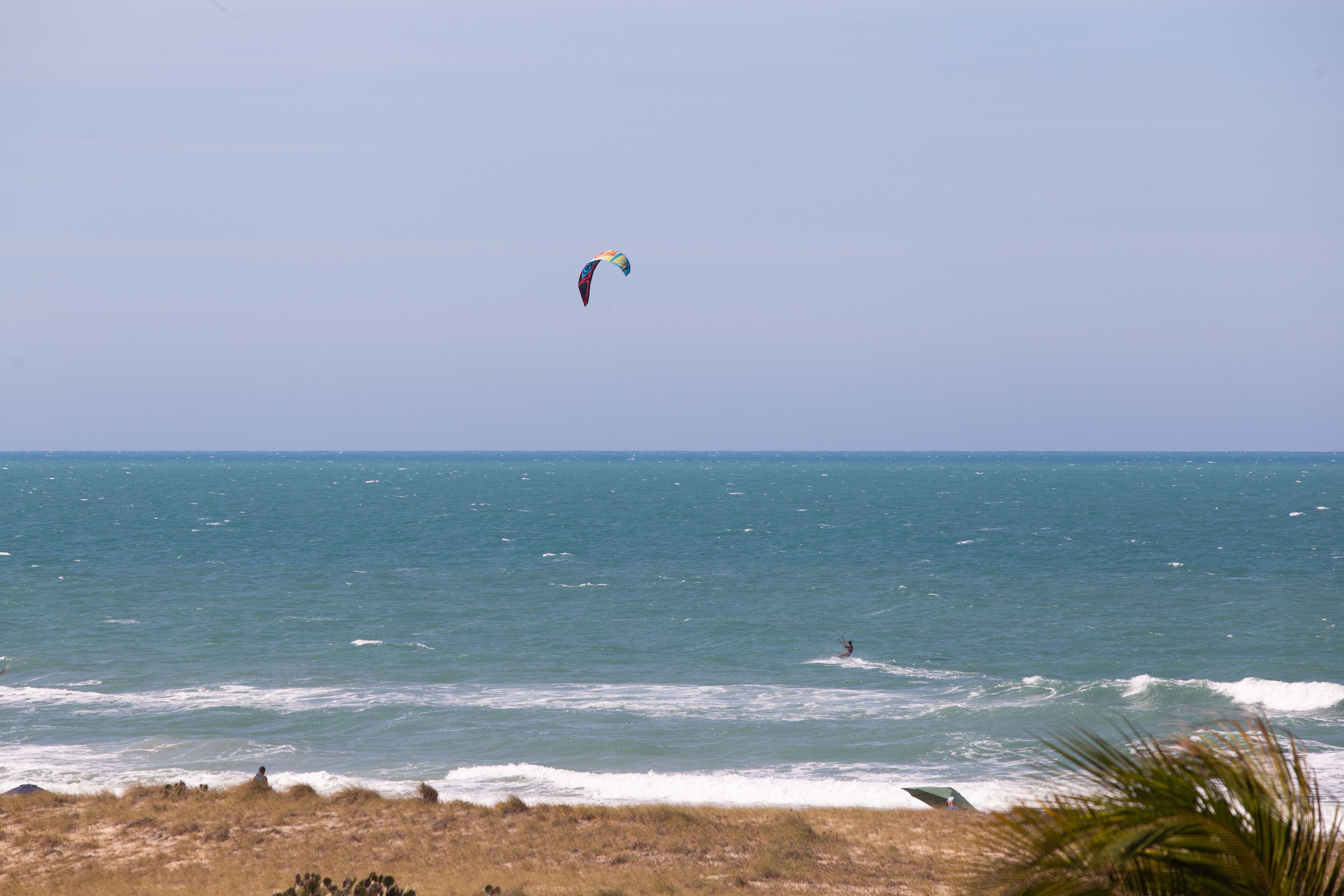 Una playa con una persona haciendo kitesurf a lo lejos.