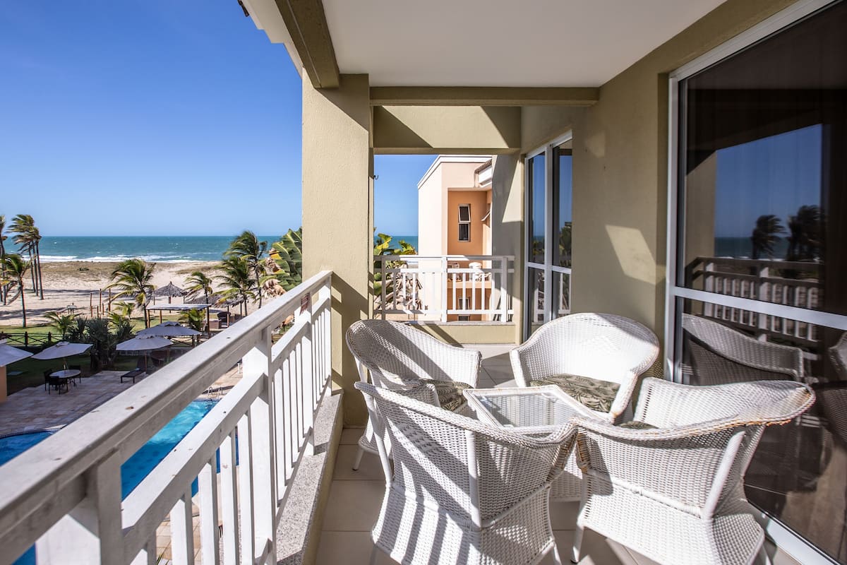 Un balcon meublé avec des meubles en osier, donnant sur une piscine et une plage.