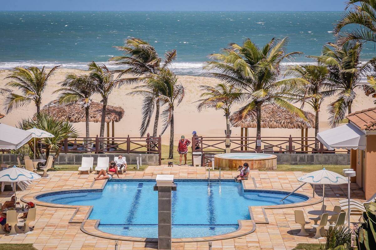 Un espace piscine offrant une vue sur la plage, les palmiers, ainsi qu’une terrasse clôturée avec des chaises longues et des parasols.