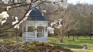 A gazebo with a white exterior and a dark roof, surrounded by trees and a grassy area.