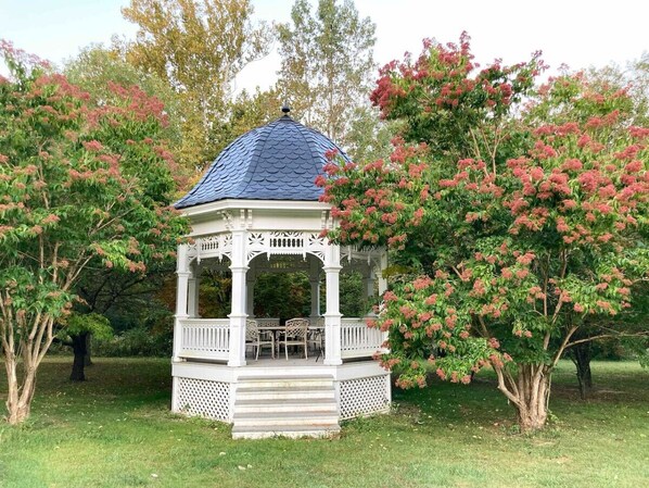 Relax in a serene garden setting, surrounded by lush greenery and flowering trees, beneath a gazebo-style structure with a seating area.