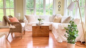 A cozy living room with a wicker chair, a white sofa, a wooden coffee table, and a potted plant.